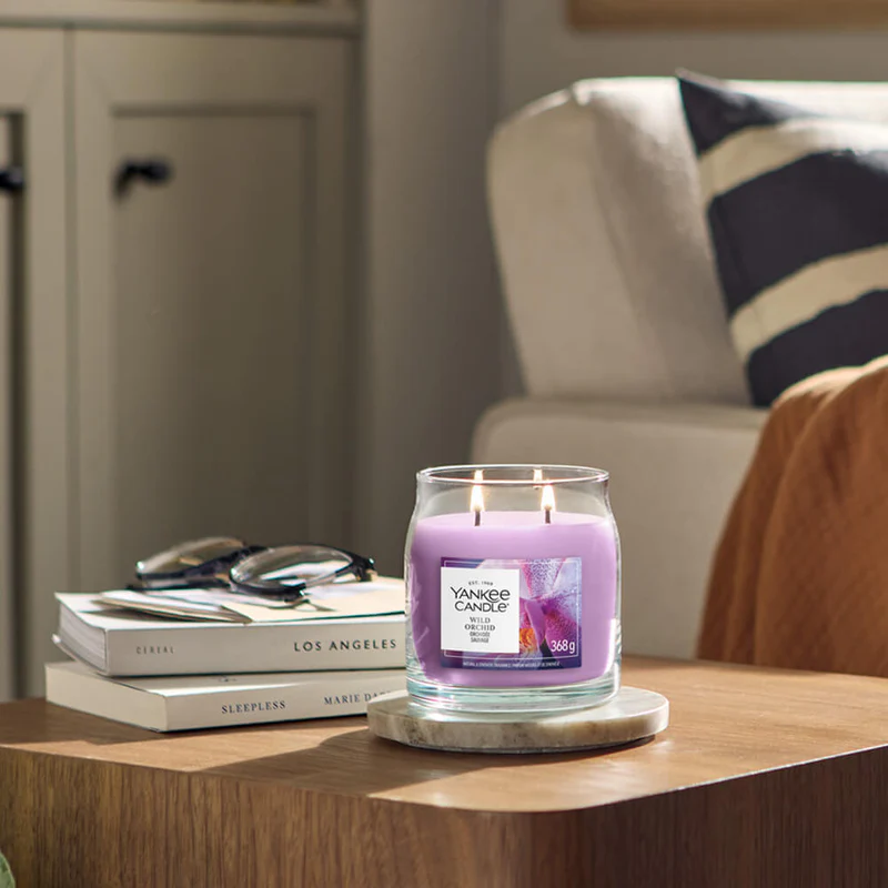 Wild Orchid – Candela Media – Yankee Candle 250 Punti 2 Una candela Yankee Wild Orchid - Candela Media con due stoppini accesi si trova su un tavolo di legno accanto a libri e bicchieri. Un divano beige con un cuscino a righe fa da sfondo, mentre il delicato profumo di orchidea selvatica riempie la stanza.