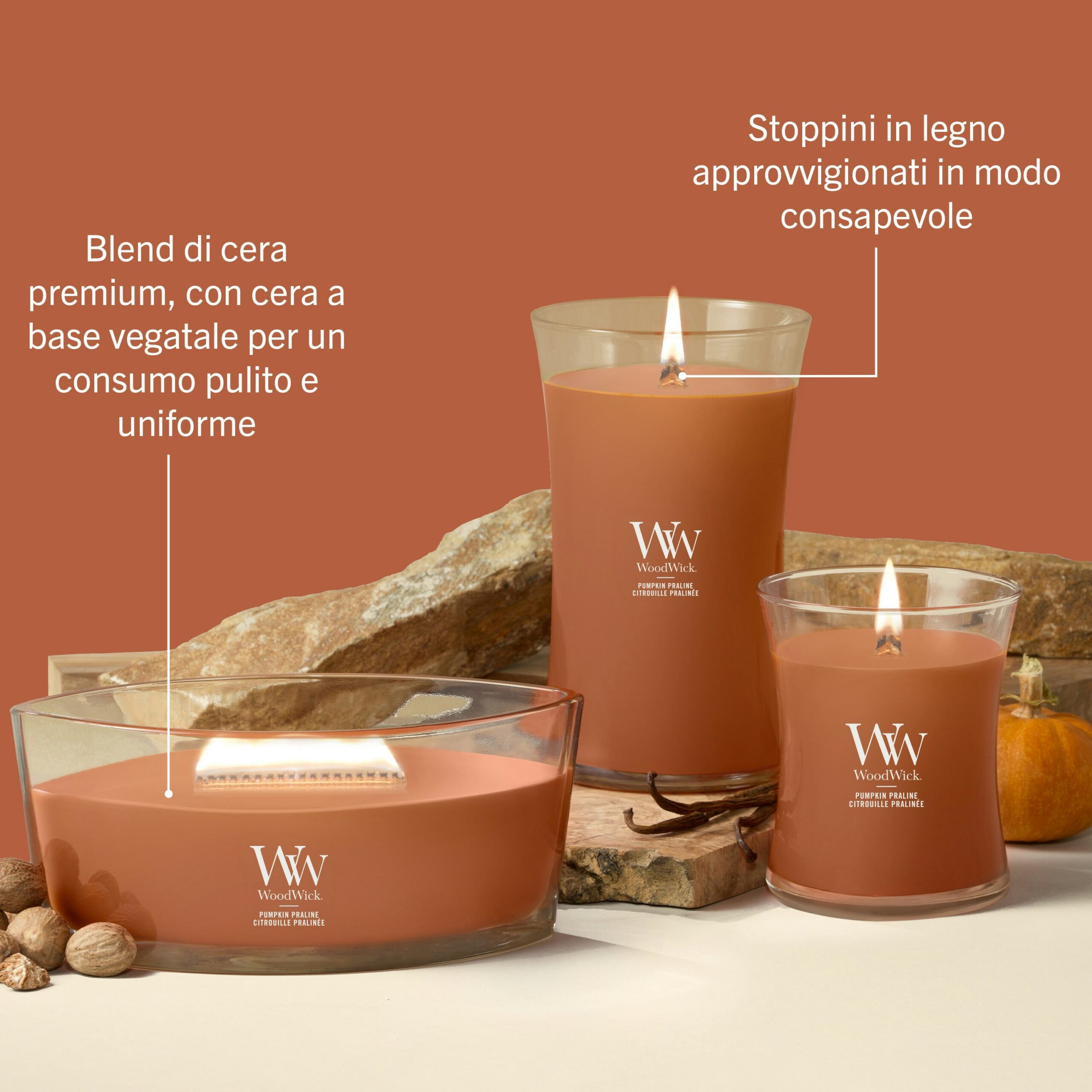 Pumpkin Praline – Candela Grande – Woodwick Giara Grande 3 Tre candele WoodWick color arancione, della linea Pumpkin Praline - Candela Grande, sono esposte in contenitori di vetro su uno sfondo beige, accanto a sassi e a una zucca. Il testo in italiano sottolinea la qualità della loro cera di soia premium e degli stoppini in legno provenienti da fonti responsabili.