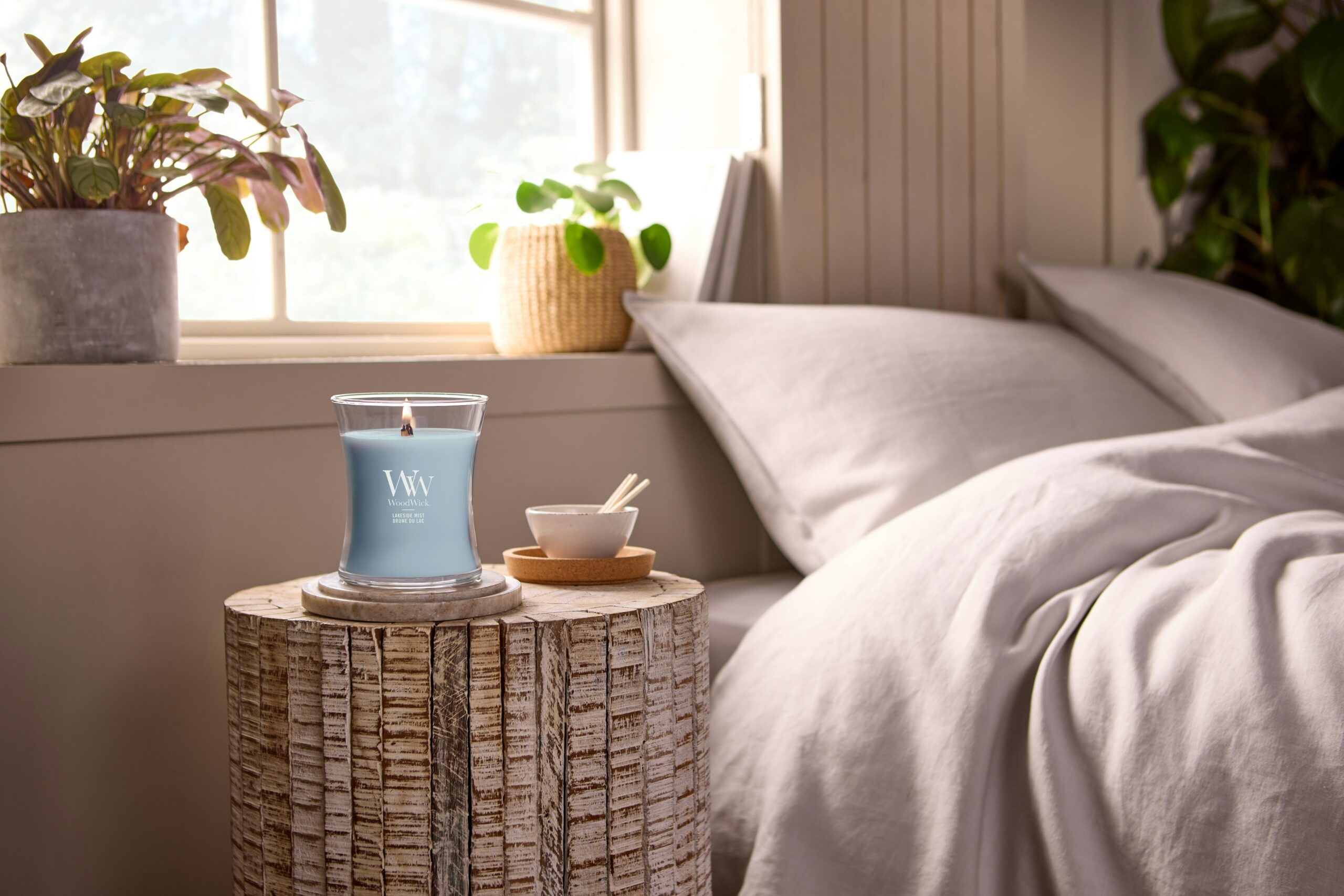 Lakeside Mist – Candela Media – Woodwick 250 Punti 3 Una candela “Lakeside Mist” - Candela Media - Woodwick accesa, una piccola ciotola con bastoncini e una pianta poggiano su un comodino di legno accanto a un letto ben rifatto con biancheria dai toni chiari, illuminato dalla morbida luce naturale che filtra da una finestra vicina. Il rilassante crepitio e il profumo rinfrescante di “Lakeside Mist” riempiono la stanza, creando un'atmosfera accogliente e tranquilla.