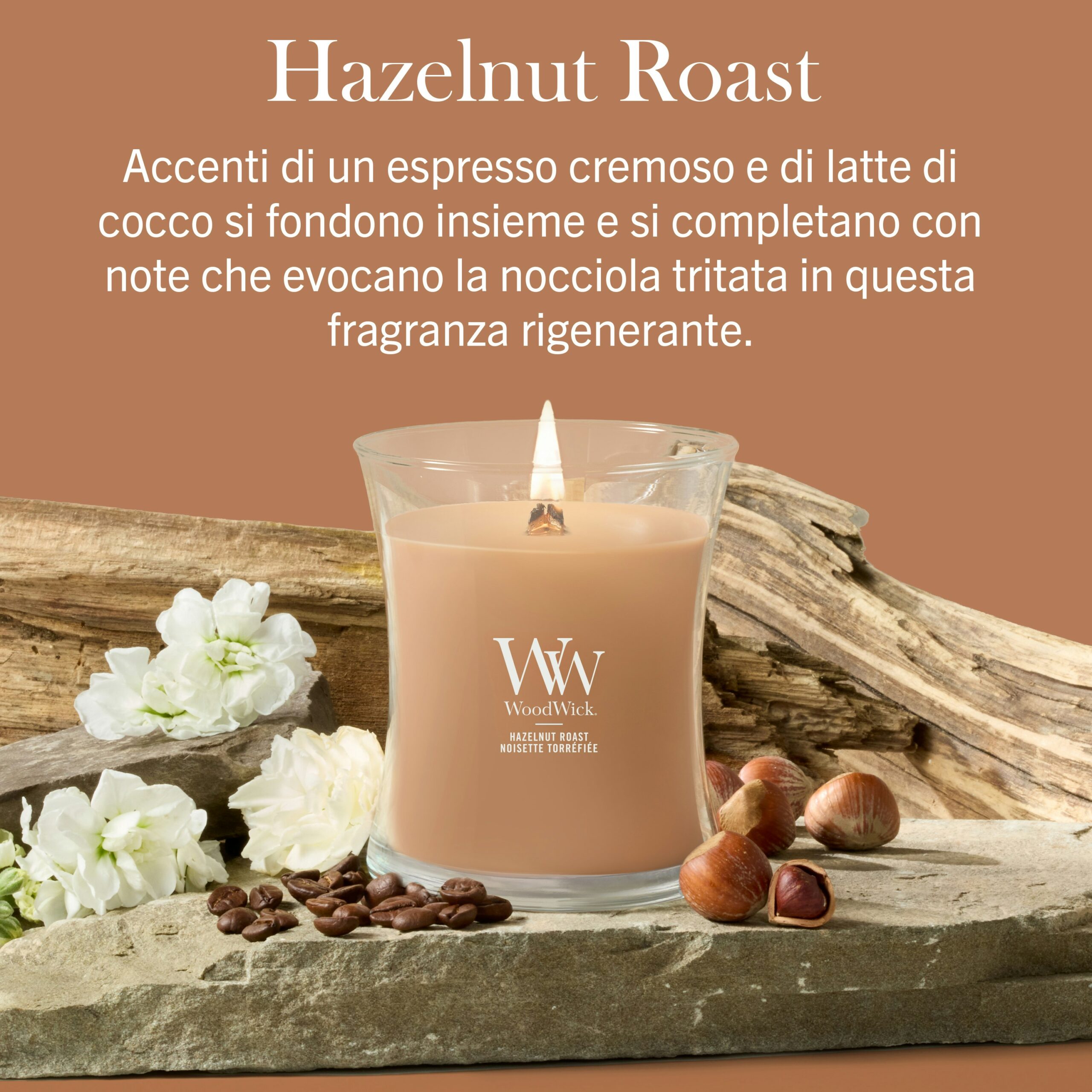 Hazelnut Roast – Candela Media – Wodwick Giara media Una miscela di nocciole tostate - Candela Media - Una candela Wodwick color beige poggia su un pezzo di legno galleggiante accanto a fiori bianchi e nocciole, su uno sfondo marrone con il testo della descrizione della fragranza in italiano.