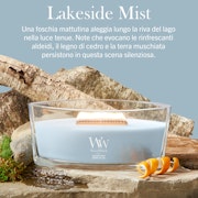 Lakeside Mist – Ellipse – Woodwick Ellipse Una candela “A Lakeside Mist - Ellipse - Woodwick”, con cera di un delicato colore blu e uno stoppino in legno, riposa tra rocce e muschio accanto a una piccola conchiglia. La scena evoca l'atmosfera serena e naturale delle rive di un lago, mentre un testo in italiano descrive la fragranza.