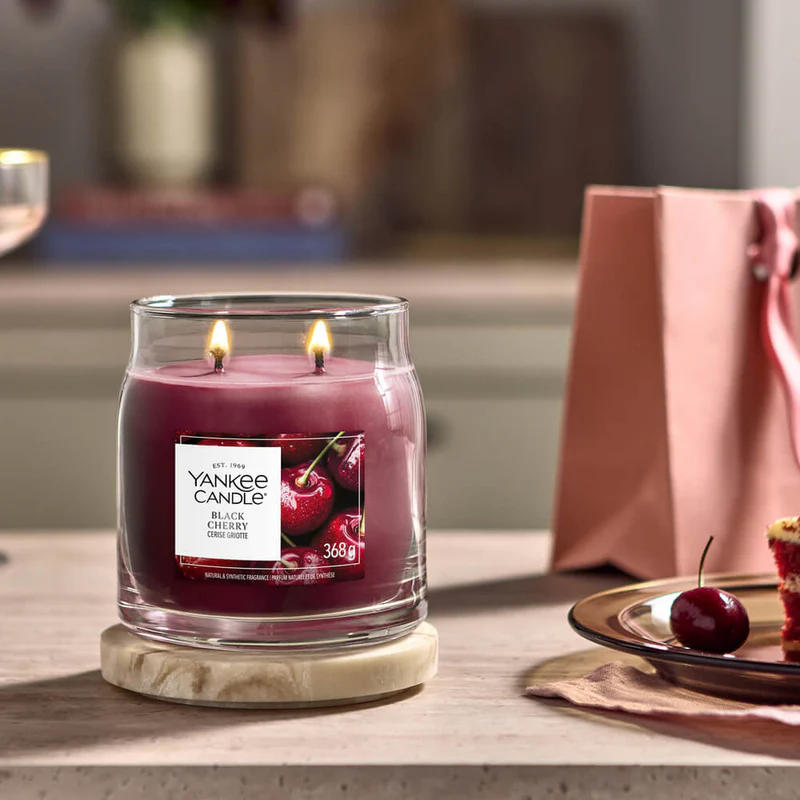Black Cherry – Candela Media – Yankee Candle 250 Punti 2 Un barattolo di Yankee Candle alla fragranza Black Cherry - Candela Media, con la candela accesa, è appoggiato su un sottobicchiere di legno. Accanto, una borsa rosa, un piatto con un dolce alle ciliegie e una ciliegia fresca su una forchetta completano l'accogliente scena sul bancone della cucina, evocando la classica atmosfera di Candela Media.