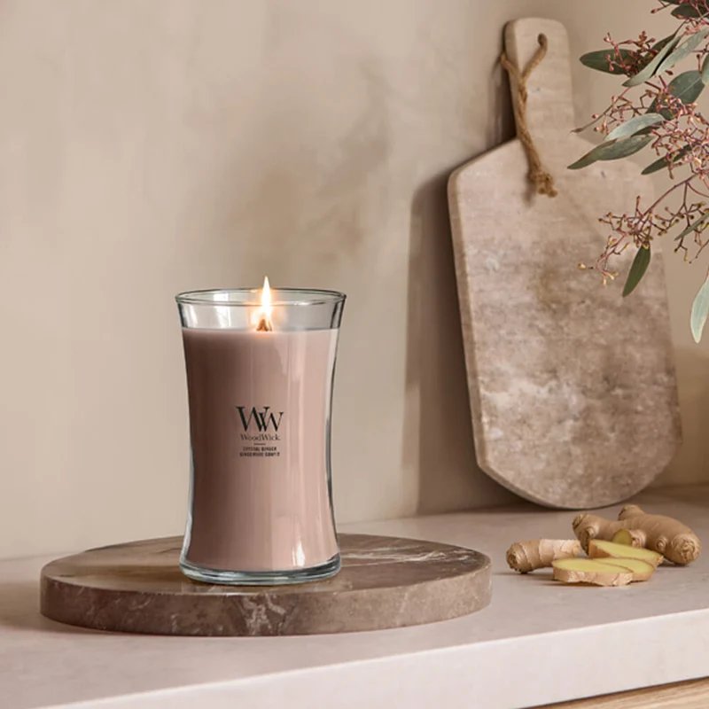 Crystal Ginger – Candela Grande – Woodwick Giara Grande 2 Una candela Crystal Ginger - Candela Grande - WoodWick accesa è appoggiata su un vassoio rotondo di marmo accanto a fette di Crystal Ginger e a un tagliere, con alcune foglie visibili ai margini dell'immagine.