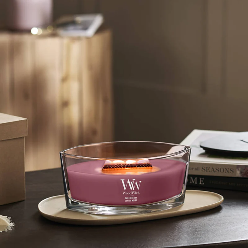 Dark Cherry – Ellipse – Woodwick Ellipse 2 Una candela WoodWick Dark Cherry Ellipse accesa, con stoppino in legno, è appoggiata su un vassoio beige posto su un tavolo, conferendo un tocco di calore a una stanza accogliente con libri e una scatola sullo sfondo.