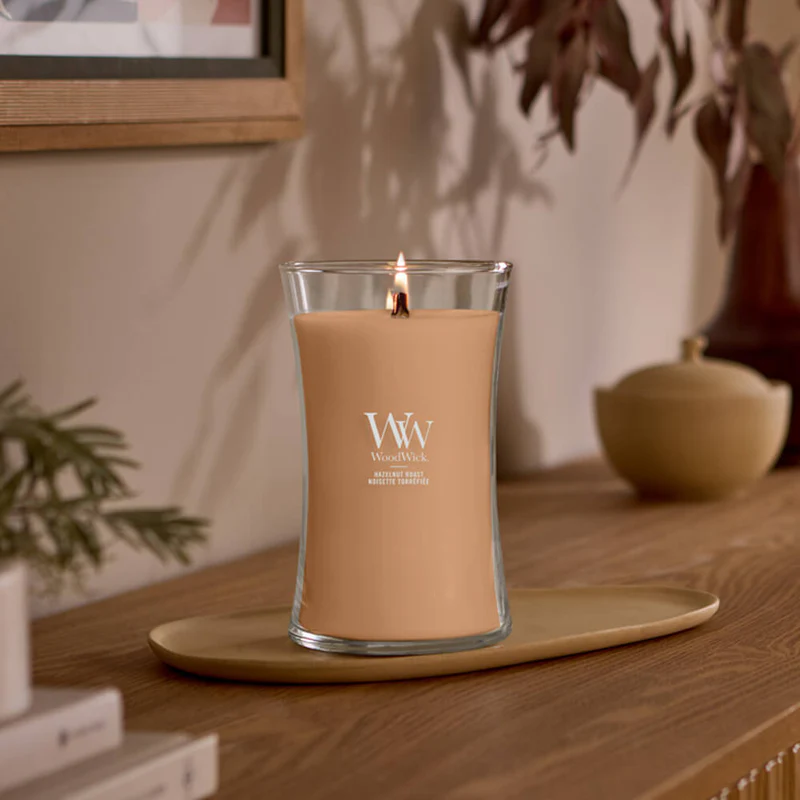 Hazelnut Roast – Candela Grande – Woodwick Giara Grande 2 Una candela “Hazelnut Roast” - Candela Grande - Woodwick, che emana una luce calda, riposa in un portacandela di vetro trasparente su un vassoio di legno, a sua volta appoggiato su una superficie di legno. L’arredamento dai toni neutri e le foglie secche si confondono delicatamente sullo sfondo.