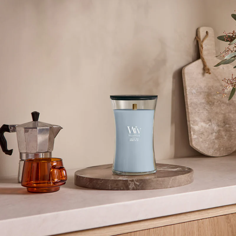 Lakeside Mist – Candela Grande – Woodwick Giara Grande 2 Una caffettiera da fornello in argento, una tazzina in vetro ambrato e una candela grigia WoodWick Candela Grande alla fragranza Lakeside Mist sono disposte sul piano di lavoro della cucina, con sullo sfondo un vassoio rotondo in pietra e un tagliere in marmo.