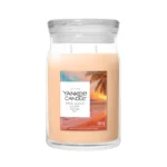 La candela "Pink Sands Glow - Candela Grande" di Yankee Candle è realizzata con cera color pesca chiaro e presenta un'immagine di un tramonto sulla spiaggia sul grande barattolo da 567 g, chiuso da un coperchio in vetro trasparente che crea un'atmosfera rilassante.