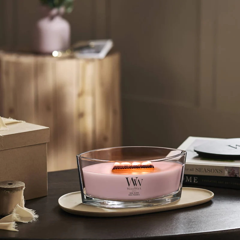 Rose Blush – Ellipse – Woodwick Ellipse 2 La candela "Rose Blush - Ellipse" di Woodwick, con le sue tre fiamme, risplende in un vaso di vetro trasparente di forma ellittica appoggiato su un vassoio beige, circondata da un arredamento morbido e accogliente sullo sfondo.