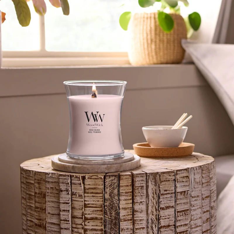 Rose Blush – Candela Media – Woodwick Giara media 2 Una candela accesa "Rose Blush" - Candela Media - Woodwick è appoggiata su un tavolino rotondo di legno vicino a una finestra, affiancata da una piccola ciotola con dei bastoncini e, sullo sfondo, da una pianta in vaso. La luce del sole inonda l'accogliente stanza.