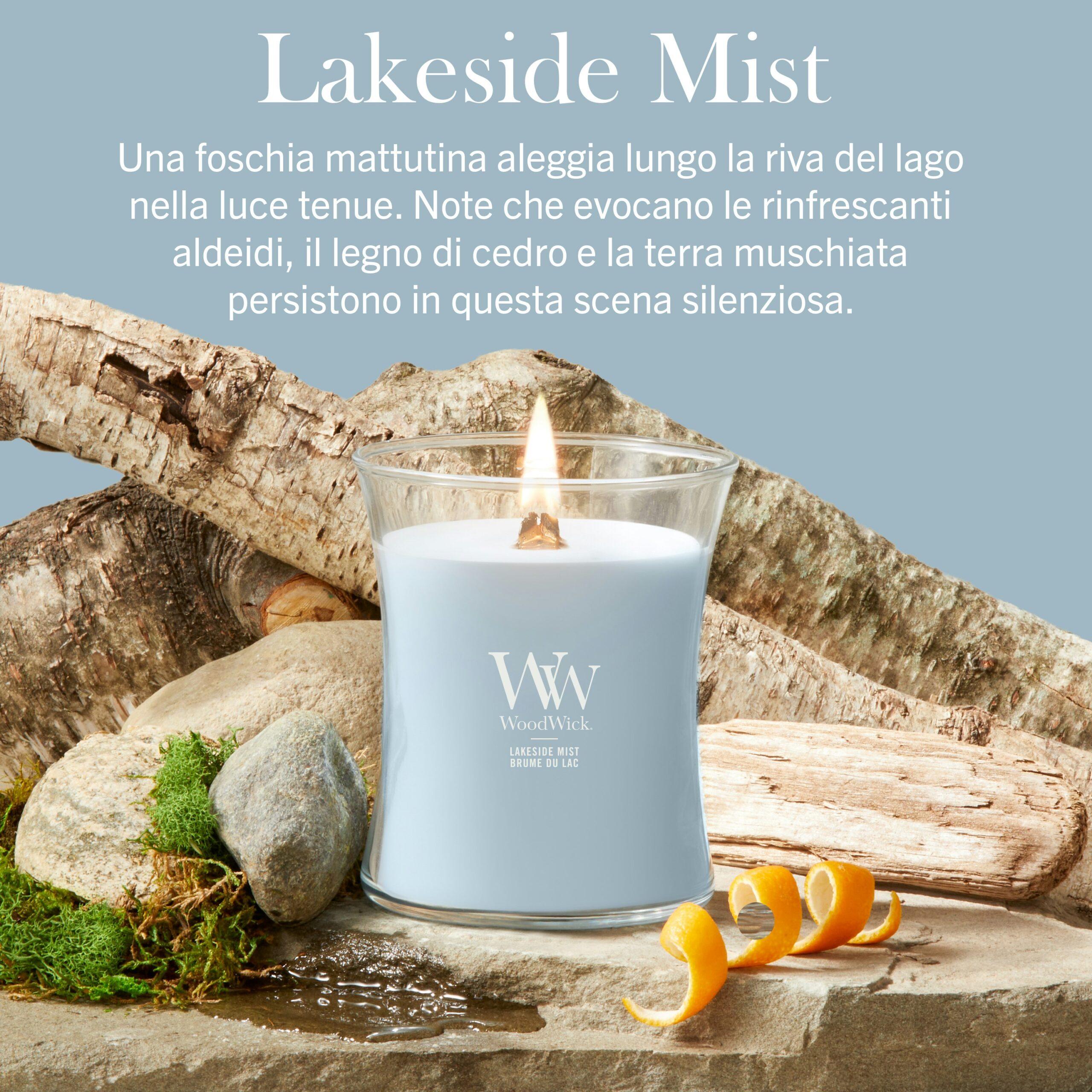 Lakeside Mist – Candela Media – Woodwick 250 Punti Una candela “Lakeside Mist” - Candela Media - Woodwick di colore azzurro chiaro riposa tra rocce e muschio, con la sua fiamma che tremola accanto a bucce d'arancia, mentre un testo in italiano descrive la sua fragranza e l'atmosfera che crea.