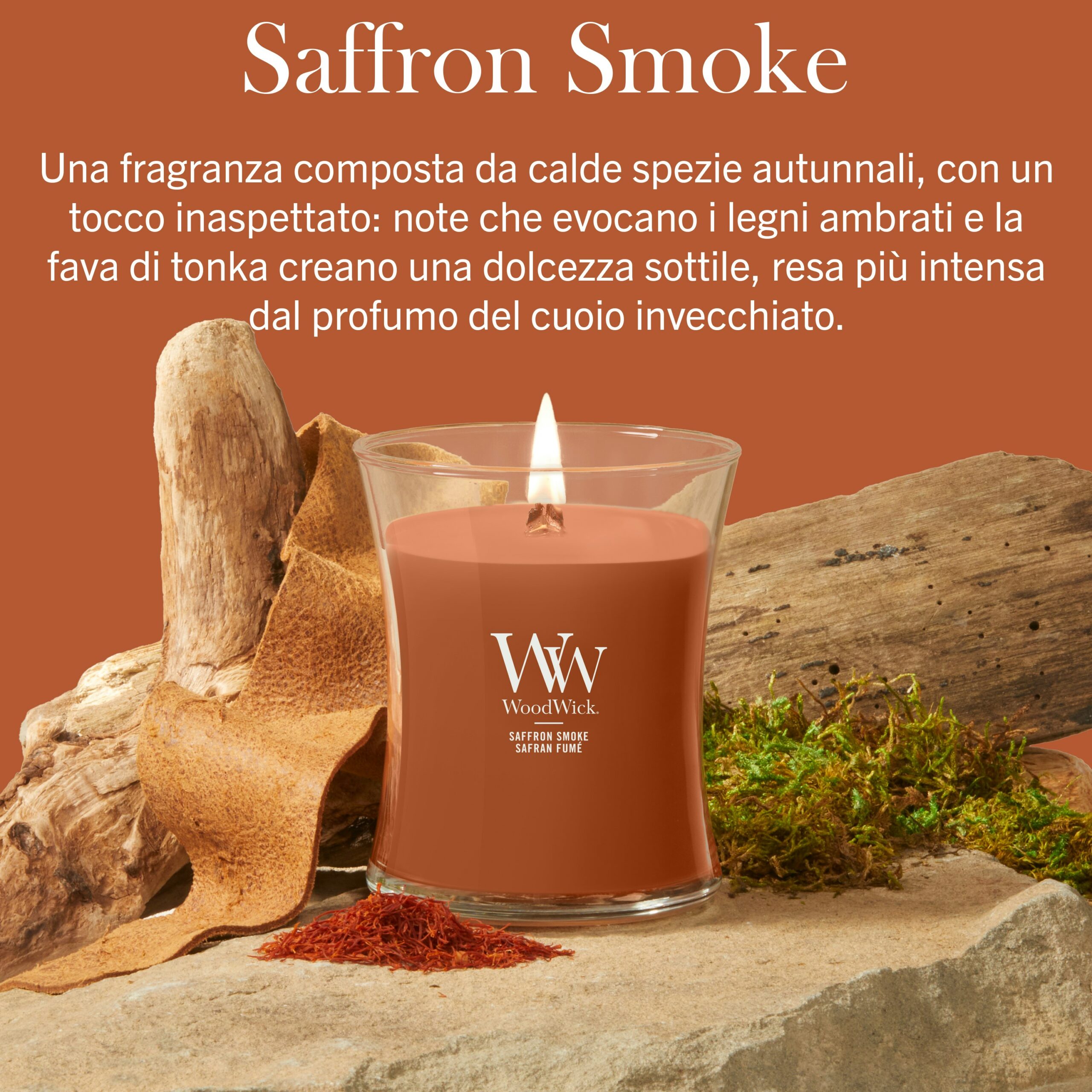Saffron Smoke – Candela Media – Woodwick Giara media A Saffron Smoke" - Candela Media - Una candela Woodwick di colore marrone poggia su una superficie beige, circondata da pezzi di corteccia, muschio e fili di zafferano. Sopra di essa, un testo in italiano ne descrive la fragranza calda, speziata e con note di cuoio.