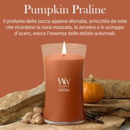 Una candela "Pumpkin Praline - Candela Grande - Woodwick" accesa è posata su un tavolo circondata da zucche, noce moscata e zenzero, con una descrizione in italiano del suo profumo autunnale. Questa grande candela riempie la stanza di note calde e accoglienti, perfette per serate intime.