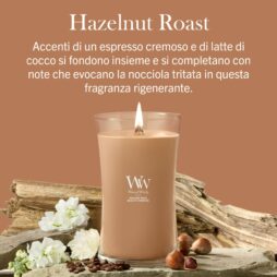 Hazelnut Roast – Candela Grande – Woodwick Giara Grande Una candela "Hazelnut Roast - Ellipse - Woodwick" di colore marrone è esposta su una base di legno, circondata da fiori e nocciole, accompagnata da un testo in italiano che ne descrive la fragranza cremosa di caffè espresso e cocco.