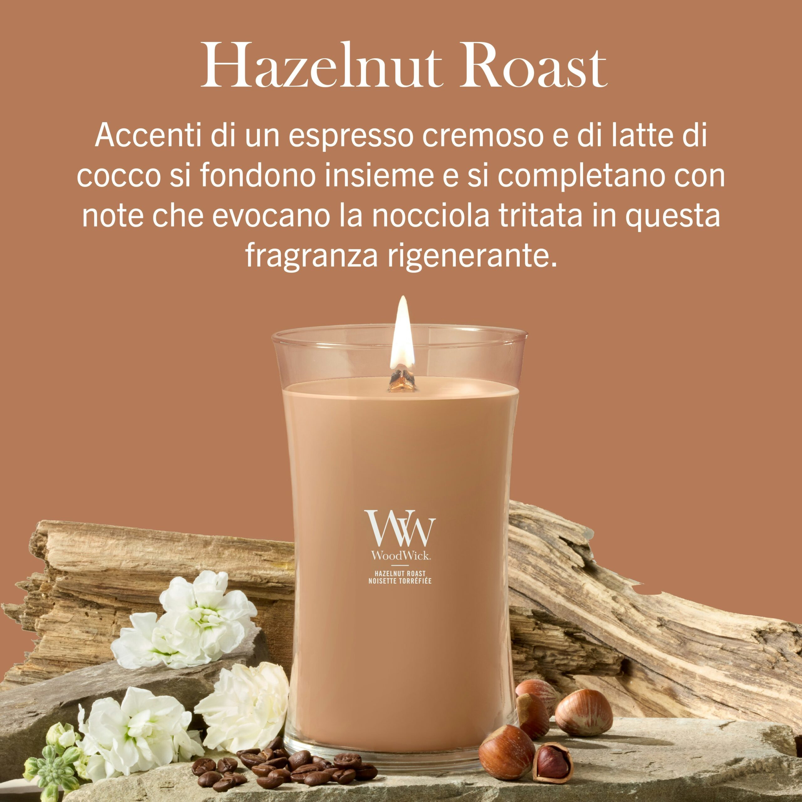 Hazelnut Roast – Candela Grande – Woodwick Giara Grande Una candela "Hazelnut Roast - Ellipse - Woodwick" di colore marrone è esposta su una base di legno, circondata da fiori e nocciole, accompagnata da un testo in italiano che ne descrive la fragranza cremosa di caffè espresso e cocco.
