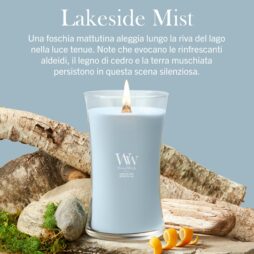 Una candela “Lakeside Mist - Candela Grande - Woodwick” accesa, di colore azzurro, è raffigurata circondata da ceppi di legno, muschio e bucce d'arancia su uno sfondo blu tenue, con un testo in italiano che descrive la fragranza.