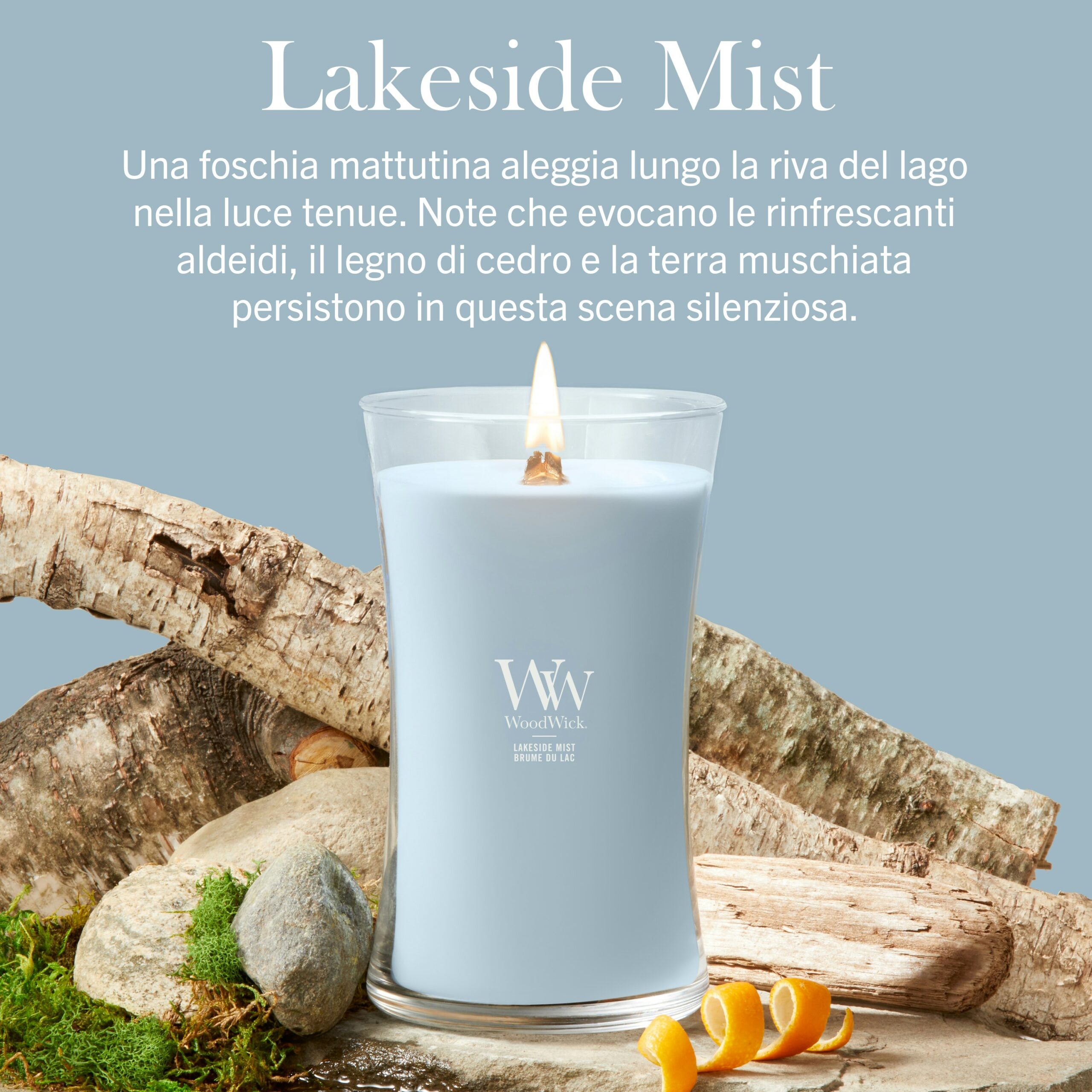 Lakeside Mist – Candela Grande – Woodwick Giara Grande Una candela “Lakeside Mist - Candela Grande - Woodwick” accesa, di colore azzurro, è raffigurata circondata da ceppi di legno, muschio e bucce d'arancia su uno sfondo blu tenue, con un testo in italiano che descrive la fragranza.