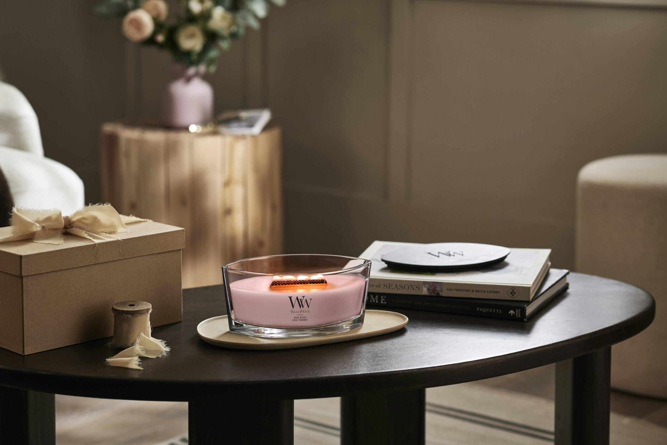 Rose Blush – Ellipse – Woodwick Ellipse 3 La candela "Rose Blush - Ellipse - Woodwick", caratterizzata da uno stoppino che emette un rilassante crepitio, risplende nel suo contenitore di vetro posato su un tavolino da caffè scuro, circondata da una pila di libri, una scatola legata con un nastro e un rocchetto, mentre un vaso di fiori aggiunge un tocco di eleganza alla scena.
