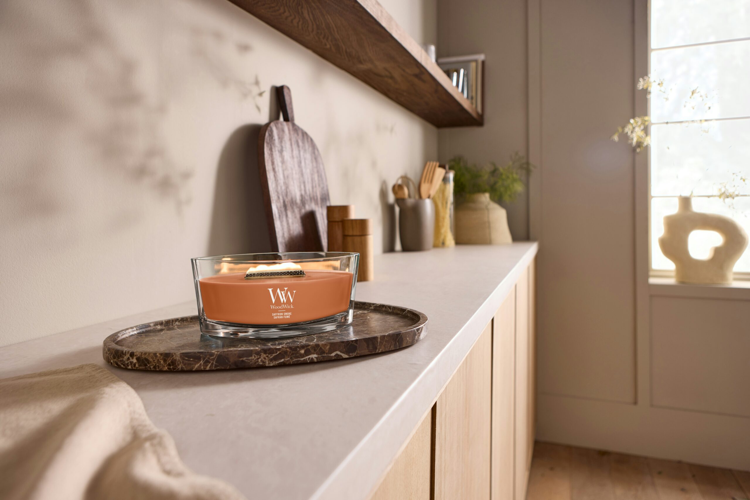 Saffron Smoke – Ellipse – WoodWick Ellipse 2 Una candela WoodWick “Saffron Smoke Ellipse” è appoggiata su un vassoio di marmo posto su un luminoso piano di lavoro della cucina, circondata da utensili di legno, barattoli e oggetti decorativi sullo sfondo, mentre la luce del sole filtra dalla finestra.
