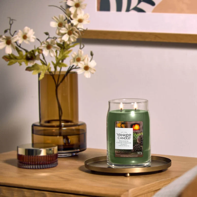 Balsam & Cedar Fireside 350 Punti 5 Una candela Yankee Candle verde, dalla invitante fragranza “Balsam & Cedar Fireside”, è accesa su un vassoio dorato accanto a un vaso di vetro marrone con fiori bianchi e a un contenitore rotondo, il tutto posato su una superficie di legno con opere d’arte appese alla parete alle spalle.