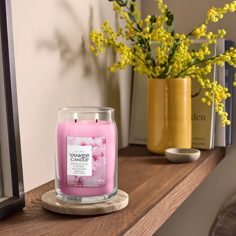 Enchanting Cherry Blossoms – Candela Grande -Yankee Candle 350 Punti 3 An Enchanting Cherry Blossoms - Candela Grande“ di Yankee Candle è appoggiata su un sottobicchiere rotondo, in cima a una mensola di legno, e diffonde nell'aria la sua delicata fragranza di fiori di ciliegio. Sullo sfondo, un vaso giallo pieno di fiori gialli e diversi libri contribuiscono a creare un'atmosfera calda e accogliente.
