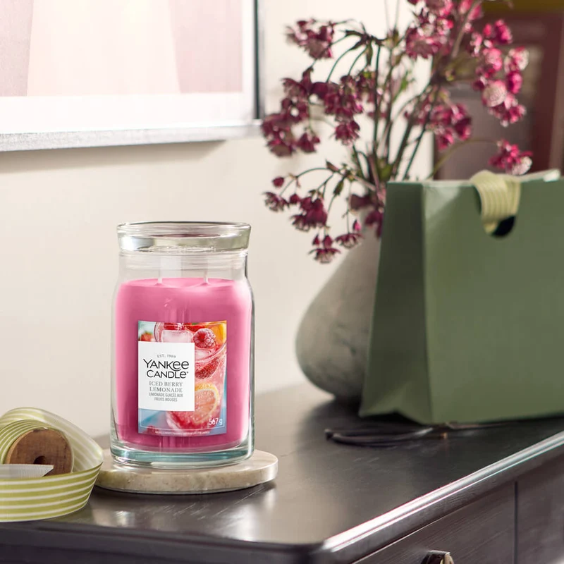 Iced Berry Lemonade – Candela Grande – Yankee Candle 350 Punti 6 Una candela “Iced Berry Lemonade - Candela Grande” della Yankee Candle, di colore rosa, è appoggiata su un tavolo di legno accanto a un sacchetto regalo verde, un vaso grigio pieno di fiori rosa, dei nastri e una foto incorniciata appesa alla parete.