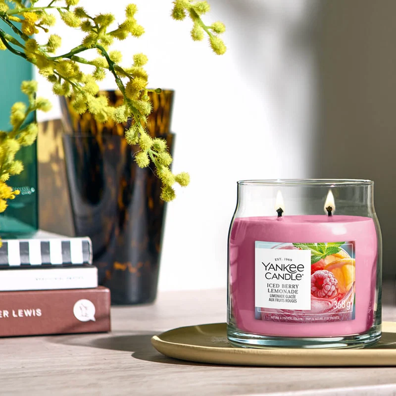 Iced Berry Lemonade – Candela Media – Yankee Candle 250 Punti 2 Una limonata ghiacciata ai frutti di bosco - Candela Media - Una candela Yankee Candle rosa è appoggiata su un piatto beige accanto a un vaso di fiori gialli e a un bicchiere di vetro marrone, creando un invitante centrotavola.