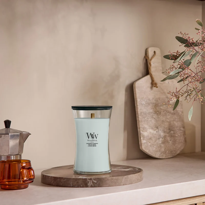 Sea Spray – Candela Grande – Woodwick Giara Grande 3 La candela “Sea Spray - Candela Grande” di Woodwick, con il suo elegante design bianco, poggia su un vassoio rotondo in pietra in una cucina dai toni neutri, accanto a una macchina da caffè, una tazza di vetro, un tagliere e un vaso con delle piante. La sua delicata fragranza “Sea Spray” infonde a questo spazio sereno una freschezza invitante.