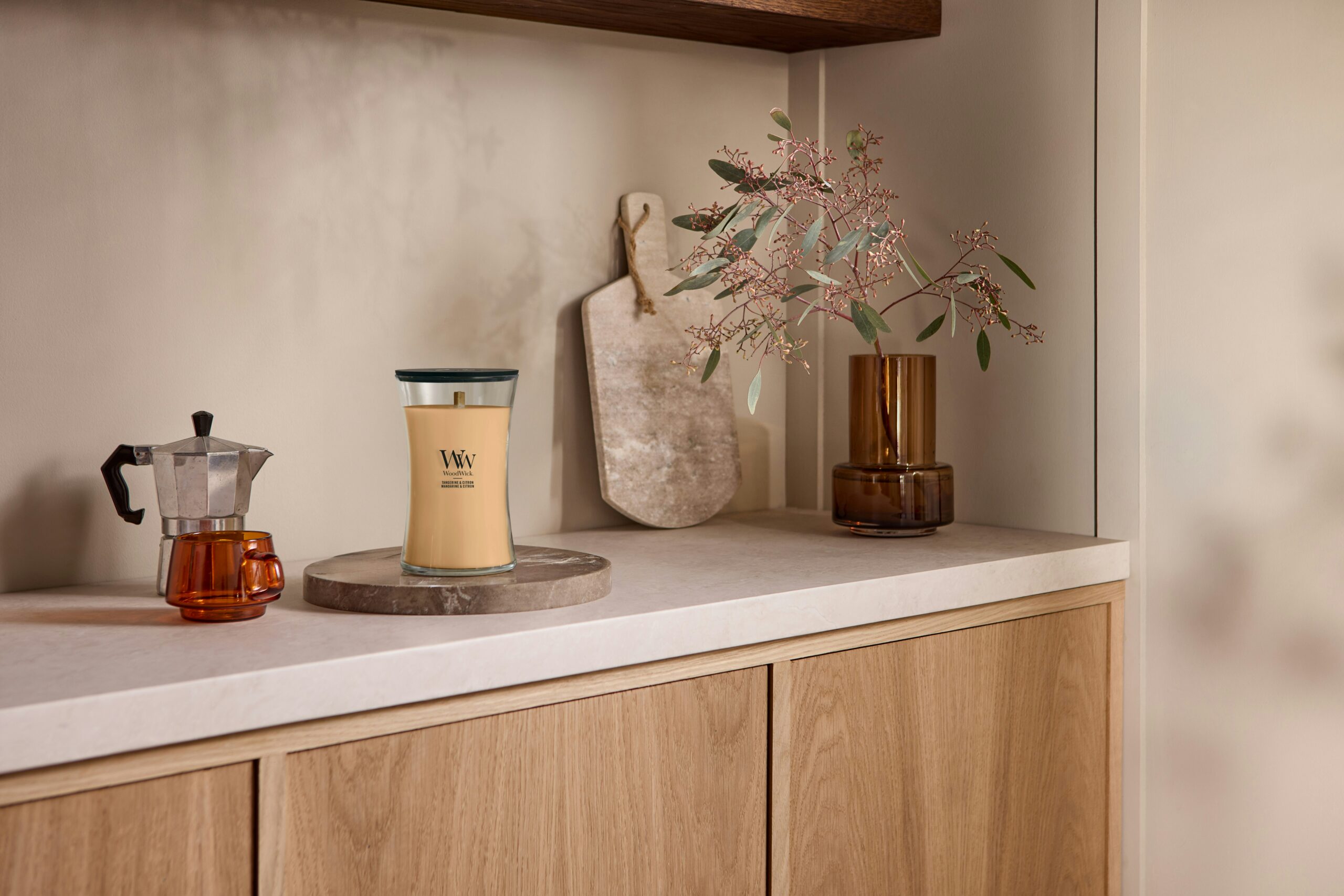 Tangerine & Citron – Candela Grande – WoodWick Giara Grande 5 Il piano di lavoro di una cucina moderna ospita una macchina da caffè, una tazza di vetro ambrato, una grande candela “Tangerine & Citrus - Candela Grande - WoodWick”, un tagliere decorativo e un vaso marrone con rami verdi. I mobili in legno chiaro e i toni neutri contribuiscono a creare un’atmosfera accogliente e minimalista, mentre il leggero profumo di agrumi della candela dona una freschezza vivace all’ambiente.