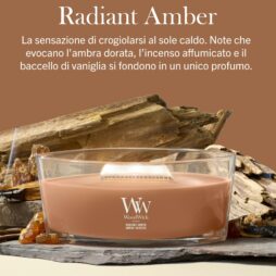 Una candela Radiant Amber - Ellipse - WoodWick con stoppino in legno, di colore marrone, è appoggiata su una superficie in pietra accanto a pietre color ambra. Sopra, un testo in italiano descrive la fragranza Radiant Amber come caratterizzata da note calde, affumicate e di vaniglia.