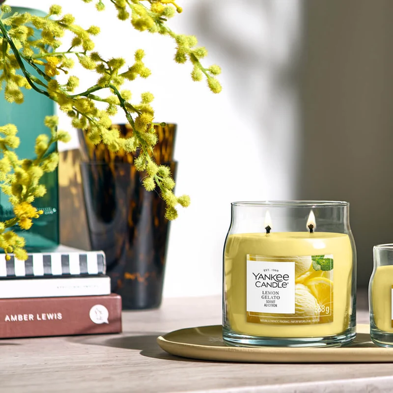 Lemon Gelato – Candela Piccola – Yankee Candle 100 Punti 2 Una candela gialla “Lemon Gelato - Candela Media - Yankee Candle” arde su un vassoio accanto a un bicchiere, alcuni libri e un vaso con fiori gialli. La luce del sole illumina questa scena accogliente, riempiendo l’aria con la rinfrescante fragranza di Lemon Gelato.
